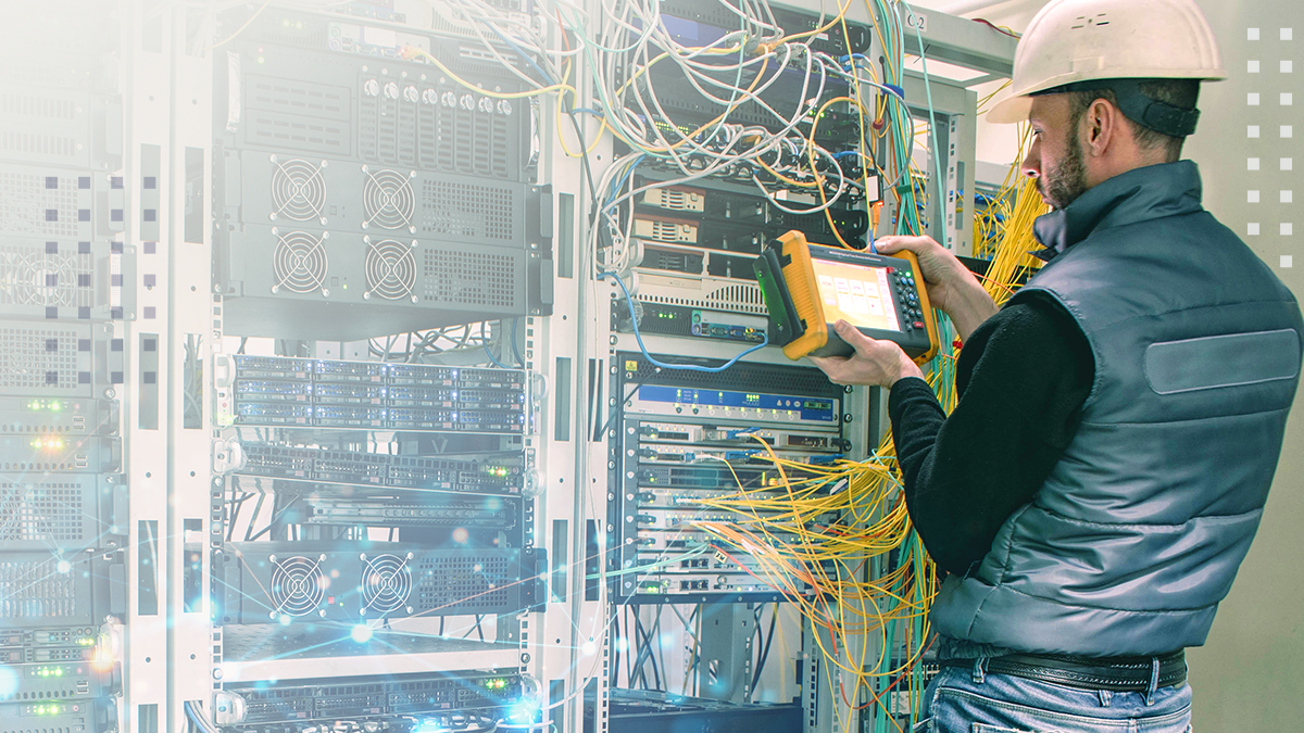 A technician wearing a hard hat operates a handheld testing device in front of a rack of network servers