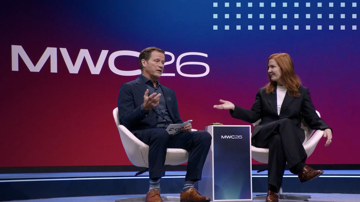 Two adults are seated on a modern stage, engaged in a discussion at the MWC26 event. The setting features a bold gradient backdrop with the large text 'MWC26' clearly visible. Both individuals are dressed in business attire, contributing to a professional and formal atmosphere. The stage design is minimalistic, with white chairs and a podium displaying the same event text.