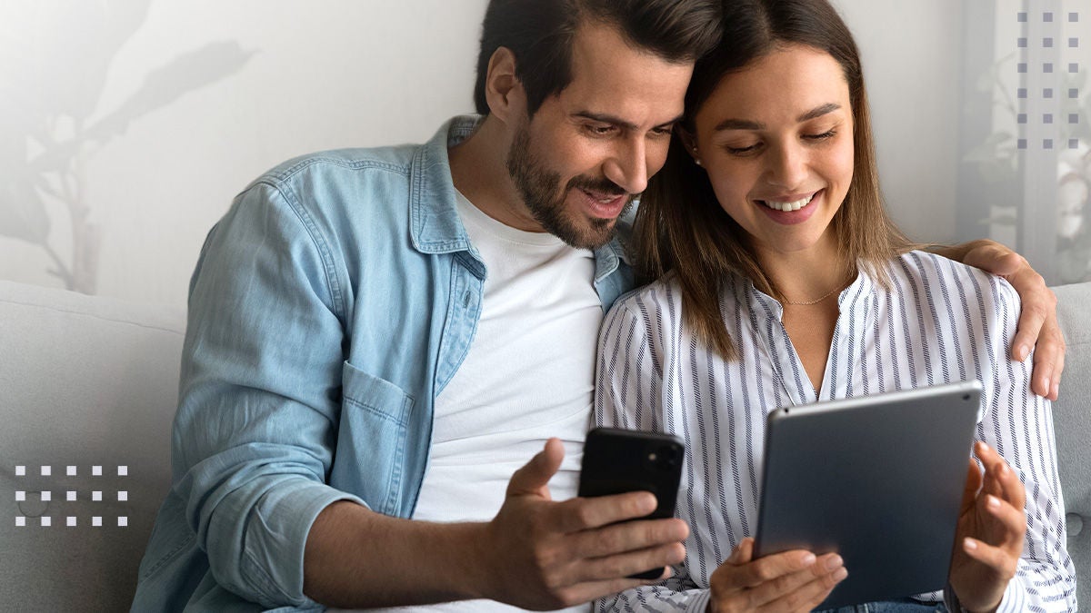 A young couple sits closely together on a light-colored sofa, each engaged with a digital device. The man holds a smartphone while the woman uses a tablet, suggesting a casual and modern home setting. Both appear relaxed, dressed in comfortable, casual clothing, with soft natural lighting enhancing the cozy atmosphere. No visible text or numbers are present in the image.