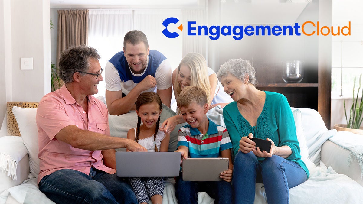 A group of adults and children sit closely on a living room sofa, interacting with laptops and a tablet