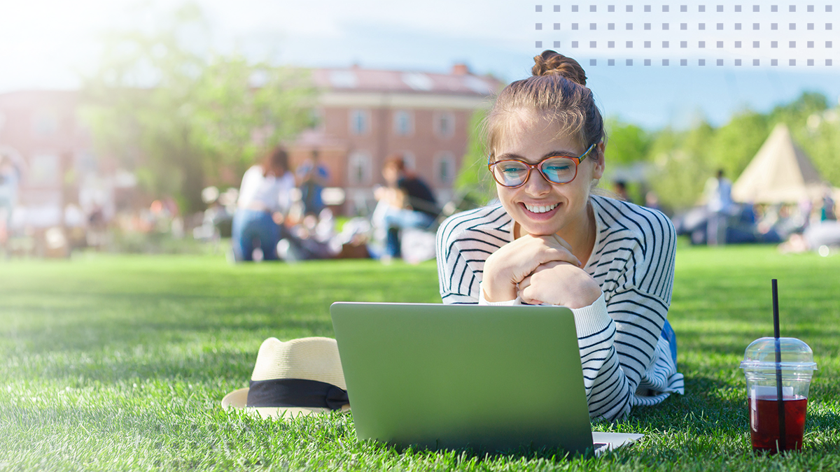 A smiling woman laying down in grass while looking at her laptop