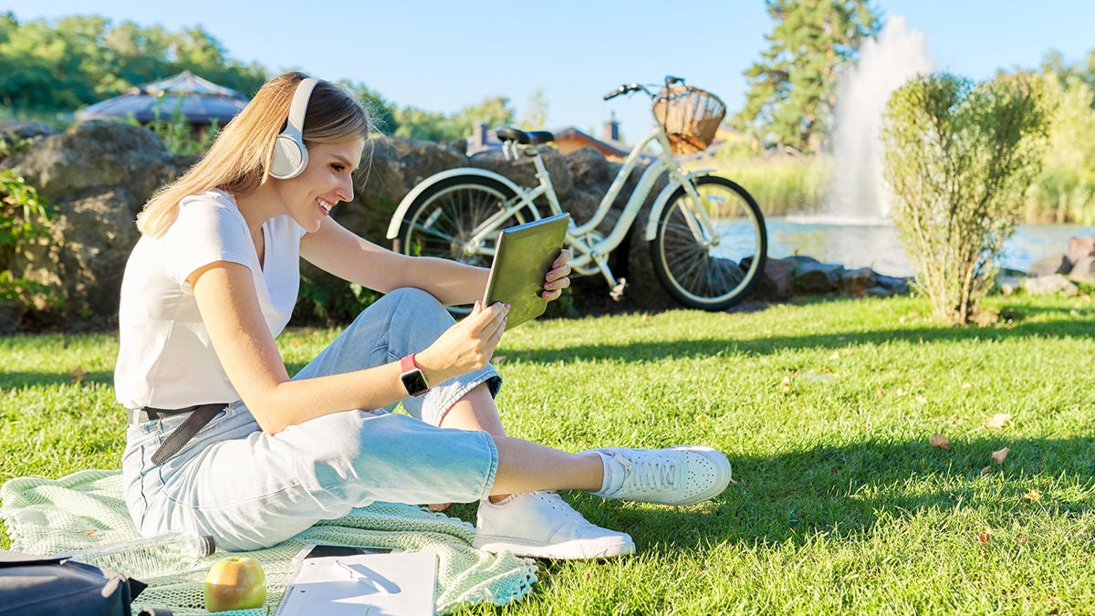 Young woman in headphones with digital tablet looking in screen of gadget, video, film, call. Female sitting on green lawn in park on sunny summer day