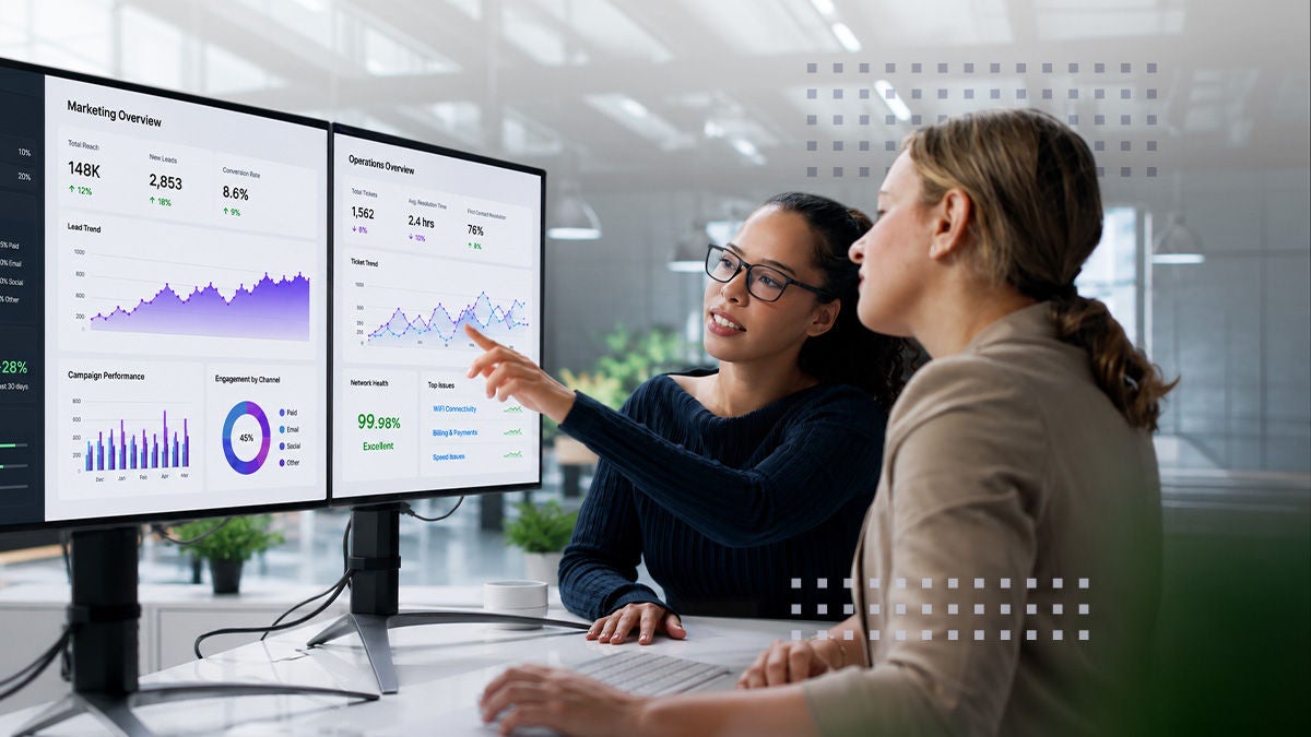 Two colleagues sit at a modern office desk reviewing marketing performance dashboards on dual monitors. One person points at a line graph while explaining data trends to the other. The screens display charts, graphs, and metrics in shades of blue, purple, and green. The background shows a bright, contemporary workspace with glass walls and a professional atmosphere.