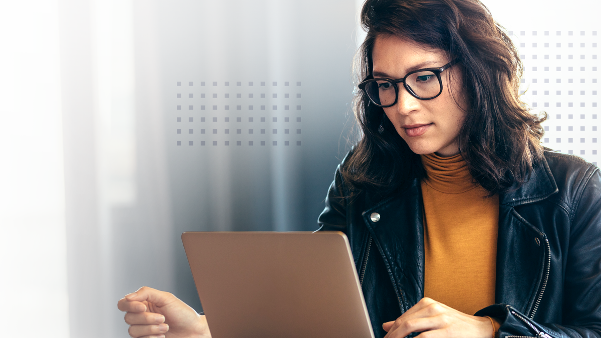 A woman wearing glasses and a leather jacket focuses on her laptop in a modern indoor setting