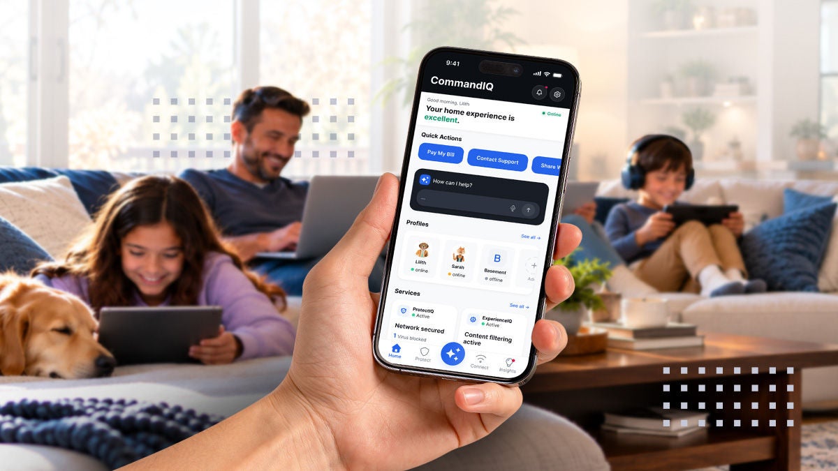 A hand holds a smartphone displaying the CommandIQ home experience app interface. In the background, a family relaxes in a cozy living room, with adults using a laptop and tablet while a child watches a device and a dog rests nearby. The scene suggests connected home management, Wi-Fi control, and digital lifestyle convenience. Soft, warm lighting and comfortable furnishings create a relaxed, modern atmosphere.