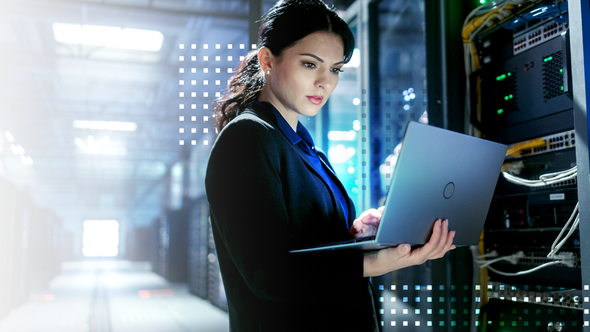 A woman stands in a data center aisle holding an open laptop while examining server equipment