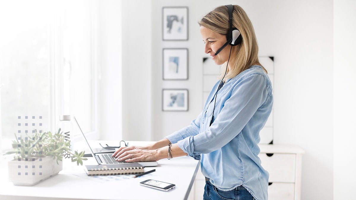 A woman stands at a white desk using a laptop and wearing headphones, suggesting remote work or virtual communication. The setting is a modern, bright home office with large windows, potted plants, and framed art on the wall. A smartphone and notebook are visible on the desk, contributing to a clean, organized workspace. The overall mood is professional and focused, with a light, airy color palette.