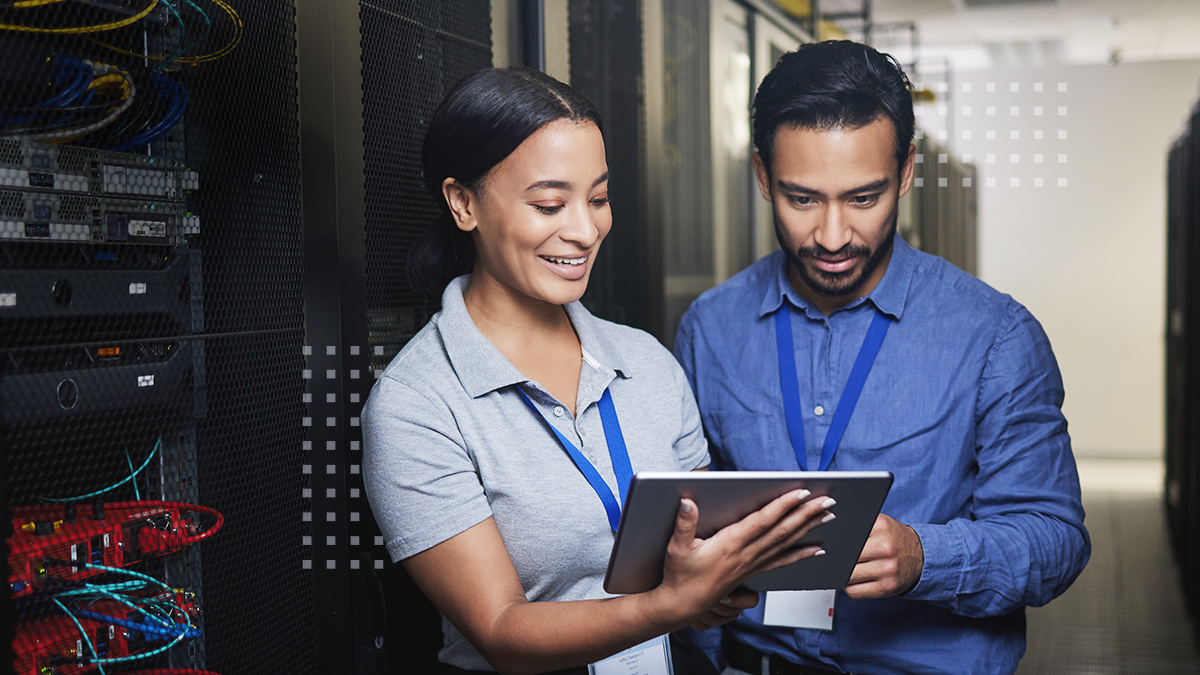Two network engineers in a server room smiling while viewing a tablet