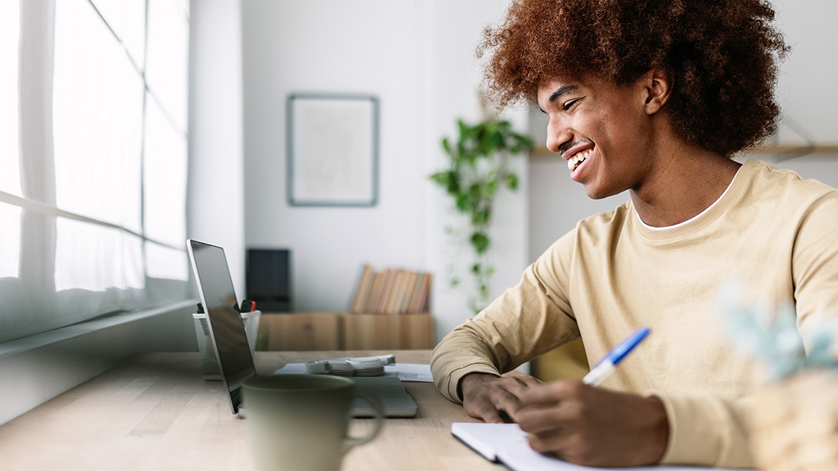 A man smiling while taking notes during an online course on his laptop